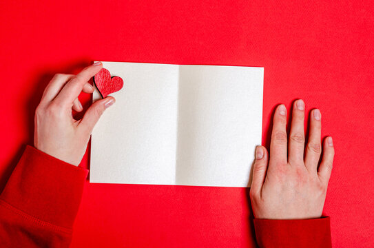 Wooden heart in female hands and a blank card on a red background for Valentine's Day