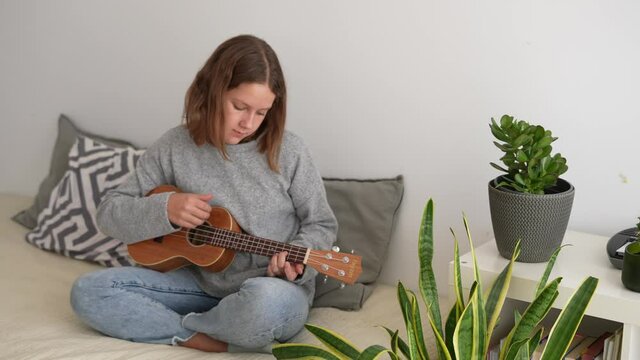 Adorable pre-teen caucasian girl playing ukulele sitting on the bed in her room. Happy and relaxed playing the songs.