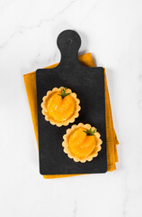 Tartlets with custard and slices of tangerines covered with syrup on a serving board on a linen napkin on a white background. Top view