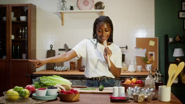 African american chef dancing in cozy kitchen, preparing ingredients for cooking vegetable salad. Woman cutting cucumber and singing. Happy housewife. Healthy vegetarian meal. 