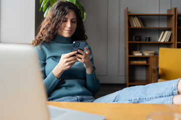 Young smiling woman freelancer relaxing at home office with mobile phone, reading internet news on...