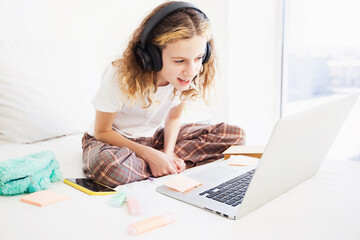 a girl child on the bed is looking at a laptop. The child is wearing headphones and waving at someone on the screen