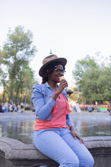 Black girl eating vanilla ice cream at a fountain in Mexico City wearing a pink blouse, denim jacket and hat