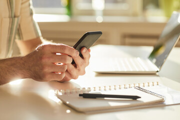 Horizontal close-up shot of unrecognizable man sitting at desk in modern office surfing Internet on smartphone