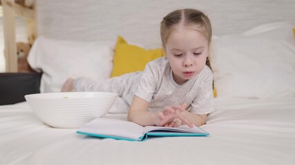 little child eats reading book while lying bed, happy family, kid daughter doing homework and eating potato chips, doing homework elementary school, first grader loves literature, children school life