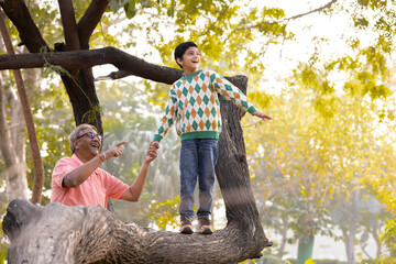 Happy grandfather holding hand of playful grandson walking on tree trunk at park
