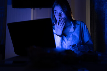 Portrait of a shocked young woman watching a horror movie at home alone at night. She covers her mouth with her hand in surprise, looking at the computer.