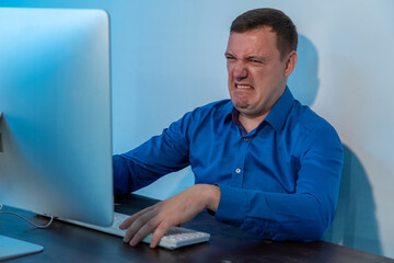 Millennial man looking disgusted in front of his computer in office. Confused businessman in blue shirt cringe and looking at screen with squeamishness at startup business office space - obrazy, fototapety, plakaty