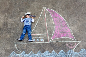 Little kid boy as pirate on ship or sailingboat picture painting with colorful chalks on asphalt. Creative leisure for children outdoors in summer. Child with captain hat and binoculars. © Irina Schmidt