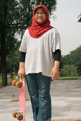 Young girl wearing hijab is running while holding pennyboard in the park. View from behind.