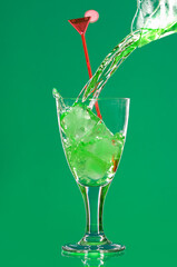 A vertical studio still life set-up of green liquid being poured in a glass with a red swizzle stick against a green background