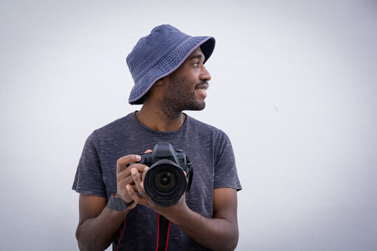 Professional photographer holds a camera in hand. Stylish and attractive man wears a bucket hat and smiles while holding a DSLR, he is isolated on white background.