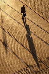 An elevated vertical view of a lady holding a bag walking across a cobbled city street at sunset, which is casting a long shadow of her body, Dresden, Germany