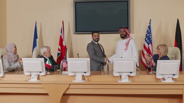 Stab shot of two foreign male politicians shaking hands at official press conference with representatives of different countries, sitting at long table equipped with computer monitors and microphones