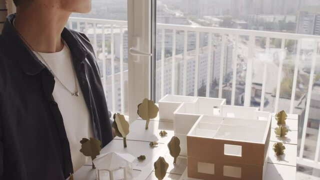 Portrait of short-haired female Asian architect posing for camera against window with big house layout in hands
