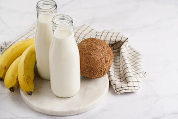 Two bottles with non-dairy milk, coconut and bananas on round marble board, grey background