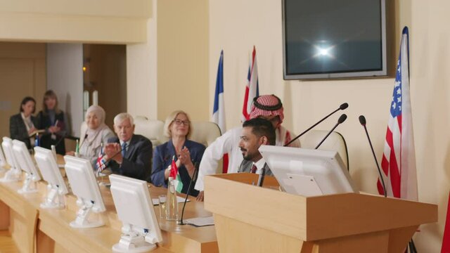 Medium shot of male politician of Arab country in traditional clothes making his speech standing on tribune of conference hall where press conference with diverse political leaders taking place