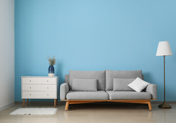 Fototapeta na wymiar Grey sofa, chest of drawers and lamp near blue wall