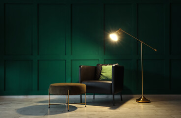 Black armchair, pouf and glowing standing lamp near green wall