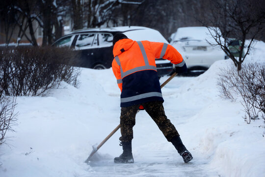 Man worker in orange uniform with a shovel cleaning the street near the car parking. Snow removal in winter city during snowstorm