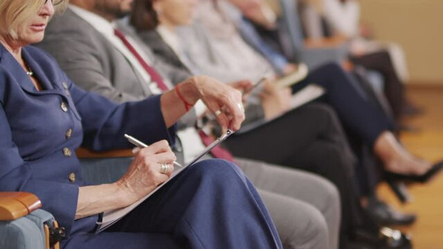 Cropped panning shot of female political leader taking notes on clipboard while listening to speaker at public conference, sitting in big auditorium with other politicians
