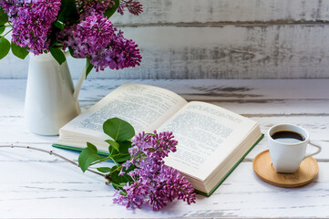 spring composition. a jug of purple lilacs, an open book with a lilac branch and a cup of fresh coffee on a white wooden table.