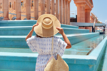 Rear view of young woman holding a wide brimmed hat and facing towards the entrance and columns of...