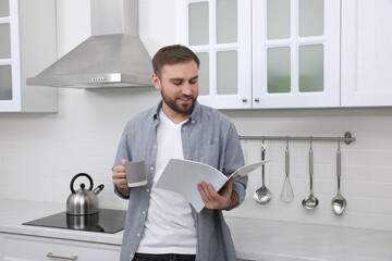 Handsome man with cup of coffee reading magazine in kitchen