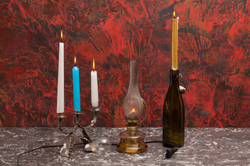 A marble top on which stands a glowing oil lamp, a candlestick with lit candles, a light bulb, a cable with a plug and a green old bottle with a lit candle. In the background is a colorful wall.