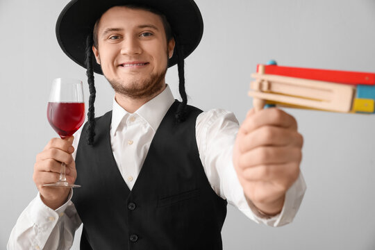 Young Jewish man with glass of wine and gragger for Purim holiday on grey background