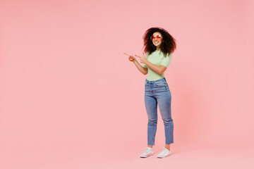 Full size body length happy young curly latin woman 20s wears casual clothes sunglasses point aside on workspace area copy space mock up isolated on plain pastel light pink background studio portrait.