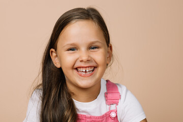 Close-up face kid with bright shining eyes and excited happy smile with teeth looking at camera wearing bright, pink jumpsuit and white t-shirt on beige background.
