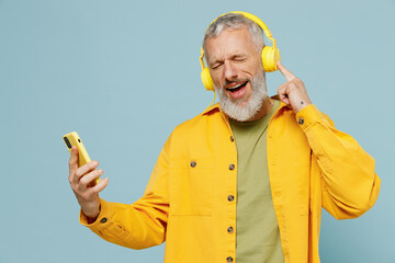 Elderly happy gray-haired mustache bearded man 50s wear yellow shirt headohones listen to music dance sing song record voice on mobile phone dictaphone isolated on plain pastel light blue background