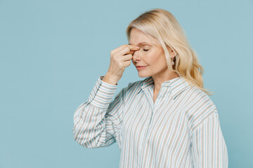 Elderly tired thoughtful caucasian woman 50s wear striped shirt keep eyes closed rub put hand on nose isolated on plain pastel light blue color background studio portrait. People lifestyle concept