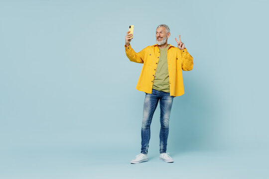 Full body elderly gray-haired mustache bearded man 50s wear yellow shirt doing selfie shot on mobile cell phone post photo on social network show v-sign isolated on plain pastel light blue background.
