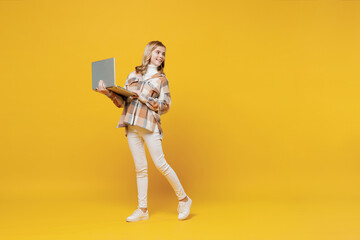 Fototapeta na wymiar Full body fun happy smiling little blonde kid girl 13-14 years wearing checkered shirt hold use work on laptop pc computer look aside on workspace isolated on plain yellow background studio portrait