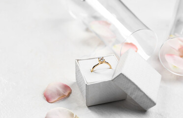 Box with golden engagement ring, glasses and rose petals on white background, closeup