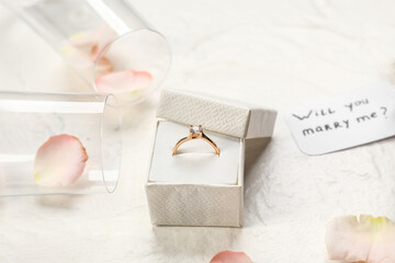 Box with golden engagement ring, glasses and rose petals on white background, closeup