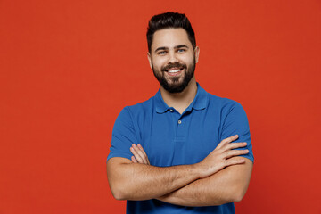 Fototapeta premium Young smiling cheerful fun cool happy caucasian attractive european man 20s in basic blue t-shirthld hands crossed folded isolated on plain orange background studio portrait. People lifestyle concept.