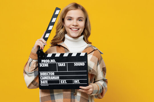 Little happy blonde caucasian kid girl 13-14 years wearing checkered shirt holding classic black film making clapperboard isolated on plain yellow background studio portrait. People lifestyle concept.