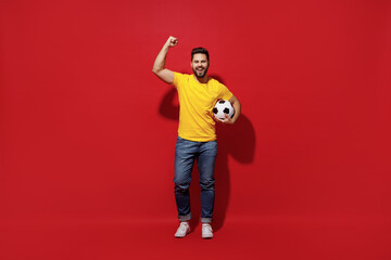 Full size body length happy young bearded man football fan in yellow t-shirt cheer up support favorite team hold soccer ball doing winner gesture isolated on plain dark red background studio portrait.