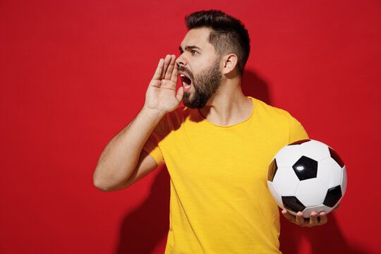Promoter angry young bearded man football fan in yellow t-shirt cheer up support favorite team hold soccer ball hold hand near mouth scream aside isolated on plain dark red background studio portrait.