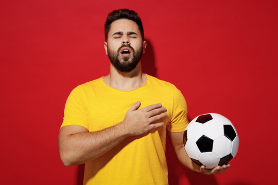 Vivid young bearded man football fan in yellow t-shirt cheer up support favorite team eyes closed hold soccer ball sing national country anthem isolated on plain dark red background studio portrait.