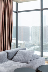 The interior of a stylish living room with panoramic windows in a city high-rise building. Gray cozy sofa and brown curtains. Vertical.