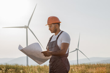 Indian technician in overalls, helmet and glasses checking blueprints of windmills while standing at rural area. Competent industrial worker controlling process of green energy production.