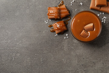Tasty salted caramel with candies in glass bowl on grey table, flat lay. Space for text - obrazy, fototapety, plakaty