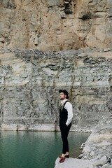 Fototapeta na wymiar Man in a suit, who is wearing a white shirt, black trousers and a black waistcoat, stands in a black hat, on a stone next to the mountains and a lake