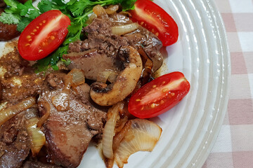 Fried chicken liver with mushrooms and onion. Fresh red tomatoes and chopped greens. Hot dish served in a white plate on a checkered rustic tablecloth