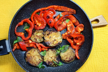 Fried sliced eggplants and sweet bell pepper with fresh basil leaves. Vegetable hot dish served in black frying pan on rustic wooden board. Yellow tablecloth. Healthy vegetarian food 