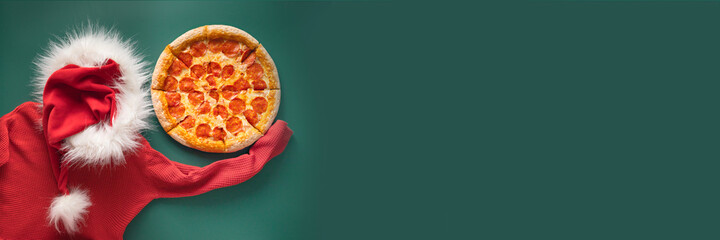 Flat lay of red santa claus hat with fluffy fur, swetear  and Pepperoni pizza on green background....